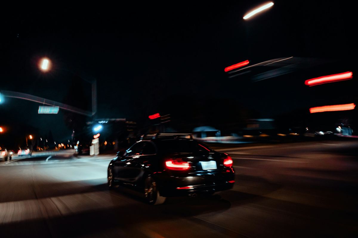 A dark sedan driving through a city at night with light trails